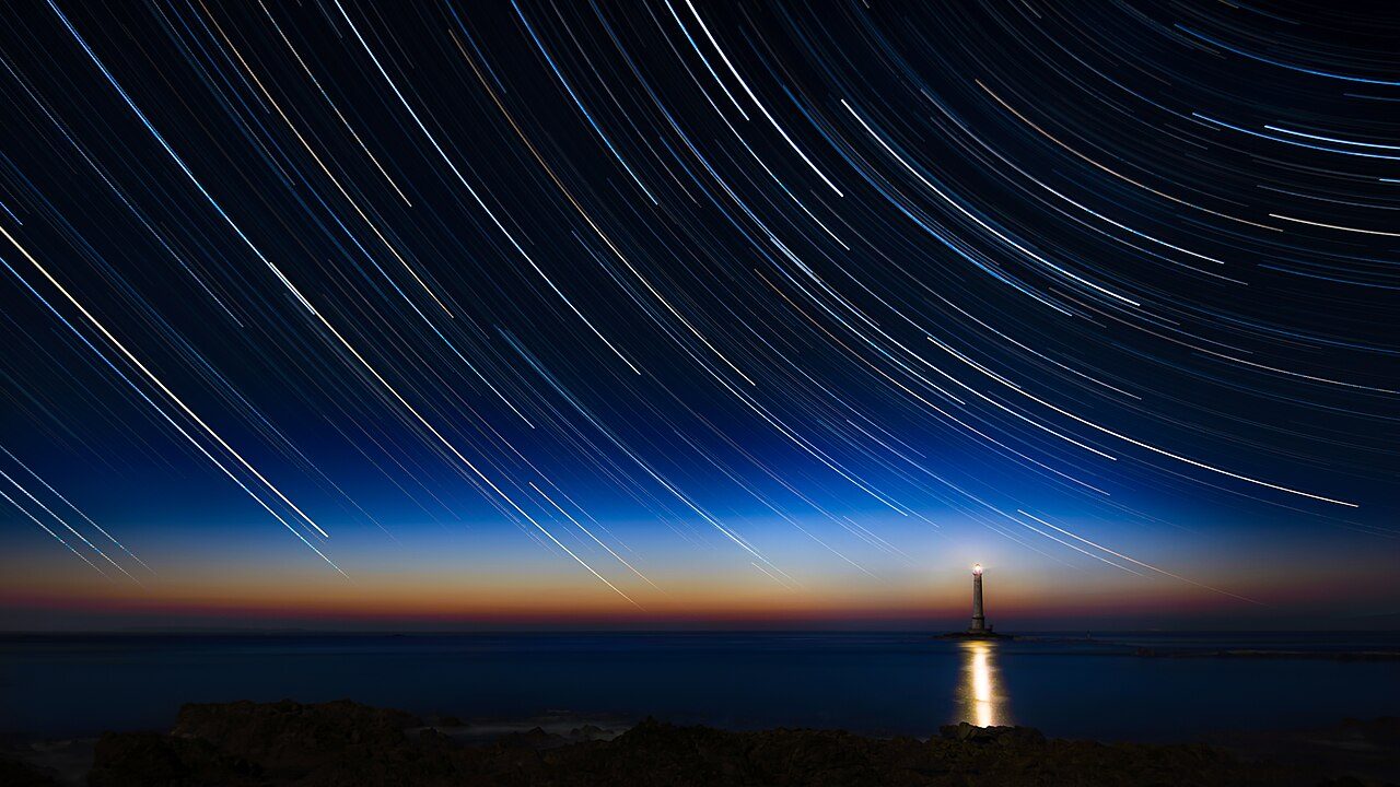 Star trails circling above a lighthouse at sunset, La Hague, France